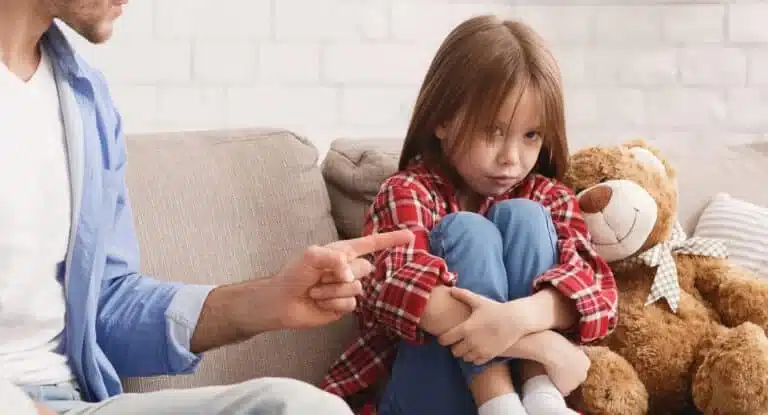 Mädchen sitzt zusammengekauert mit einem Teddybär auf dem Sofa, während ein Erwachsener mit dem Finger auf sie zeigt