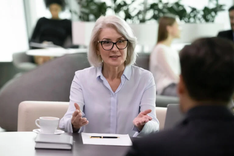 Frau mit Brille spricht in einem hellen Büro an einem Tisch mit einem Mann, der ihr gegenübersitzt