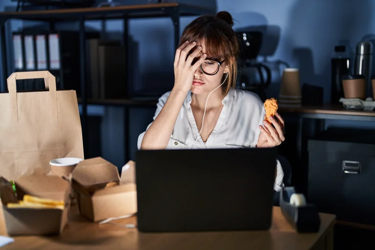 Erschöpfte Frau sitzt abends am Laptop, hält sich die Hand an die Stirn und isst Lieferessen aus Pappschachteln, Sinnbild für ungesunde Ernährung unter Dauerstress