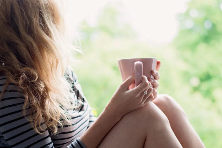Frau mit langen blonden Locken sitzt am Fenster und hält eine rosa Tasse mit beiden Händen, draußen unscharfes Grün