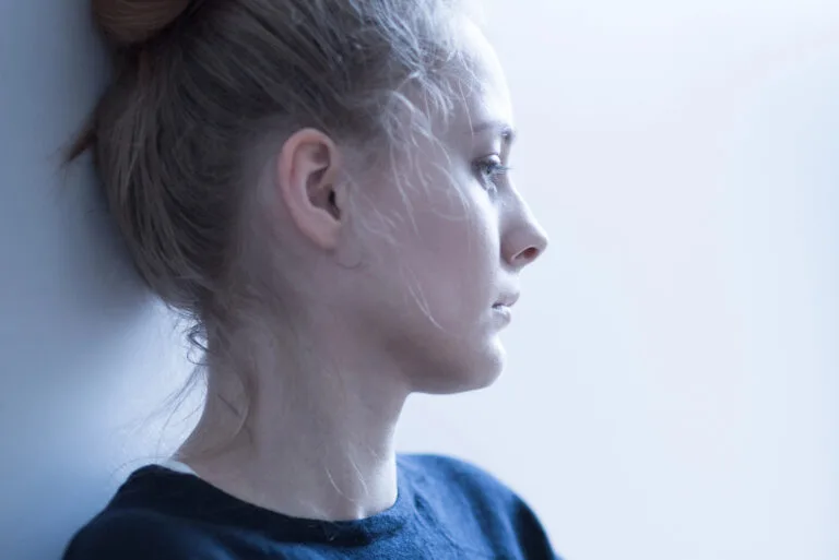 Junge Frau lehnt mit dem Kopf an einer hellen Wand und schaut nachdenklich zur Seite