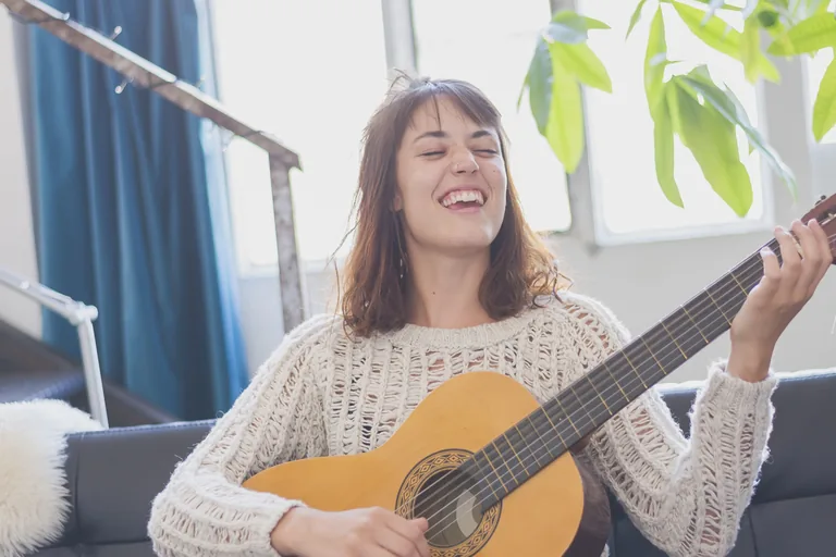 Lachende junge Frau sitzt im hellen Wohnzimmer auf dem Sofa und spielt Gitarre