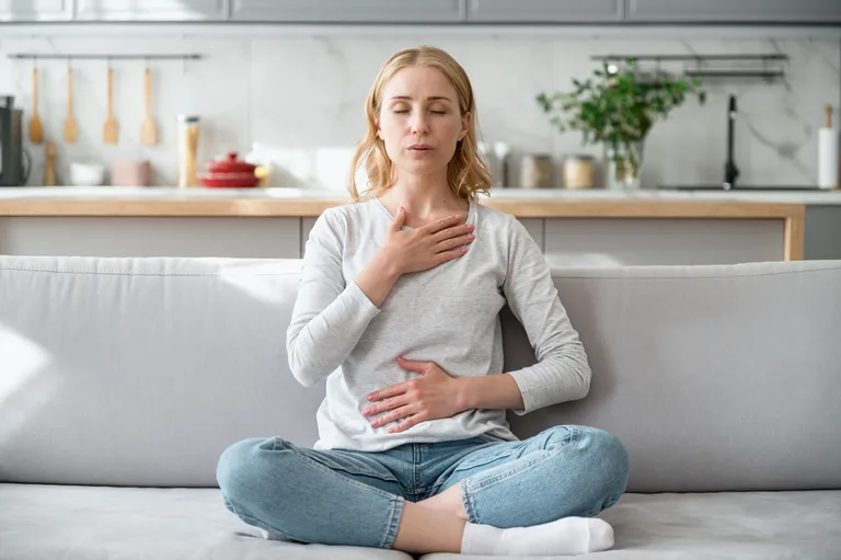 Den eigenen Körper spüren und zur Ruhe kommen 🙂 Frau sitzt im Schneidersitz auf einem hellgrauen Sofa, hat die Augen geschlossen und legt eine Hand auf die Brust und eine auf den Bauch.