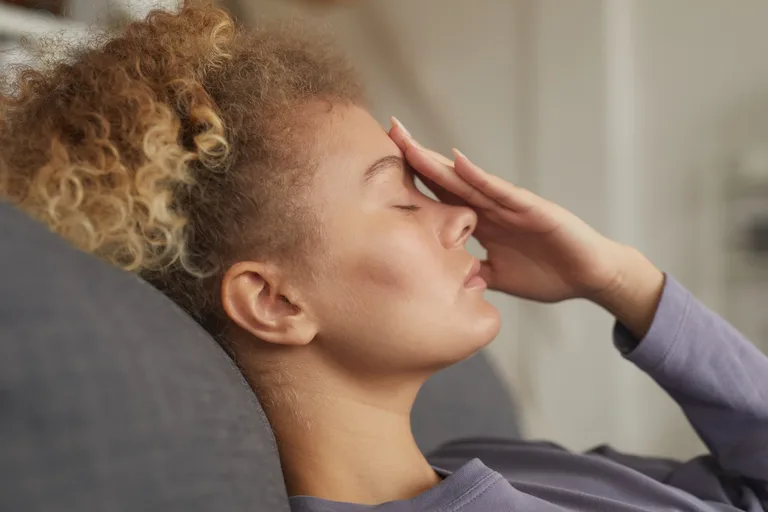 Erst einmal durchatmen und dem Körper Ruhe gönnen 🙂 Frau mit lockigem Haar lehnt mit geschlossenen Augen auf einem grauen Sofa und hält sich eine Hand an die Stirn.