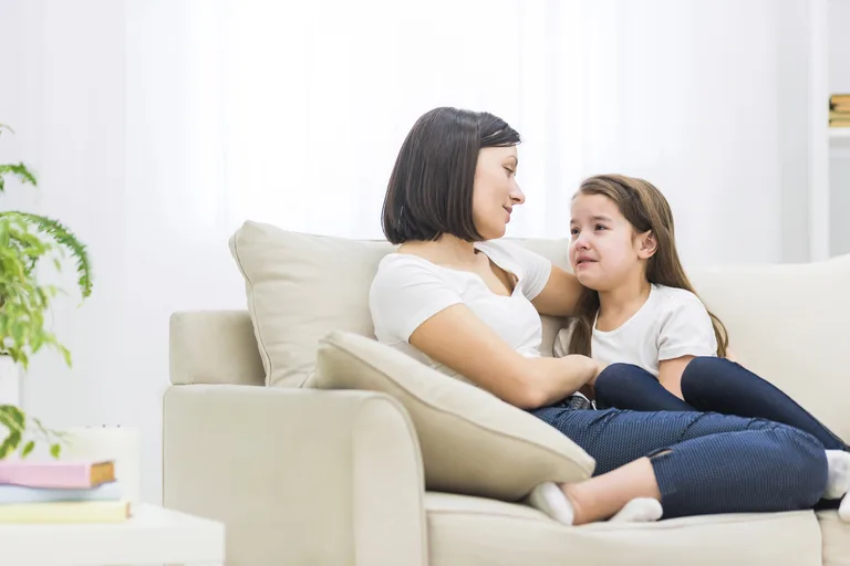 Gesehen werden gibt Kindern Halt 🙂 Mutter sitzt mit ihrer Tochter auf einem hellen Sofa, legt den Arm um sie und hört ihr zu, das Mädchen wirkt traurig.