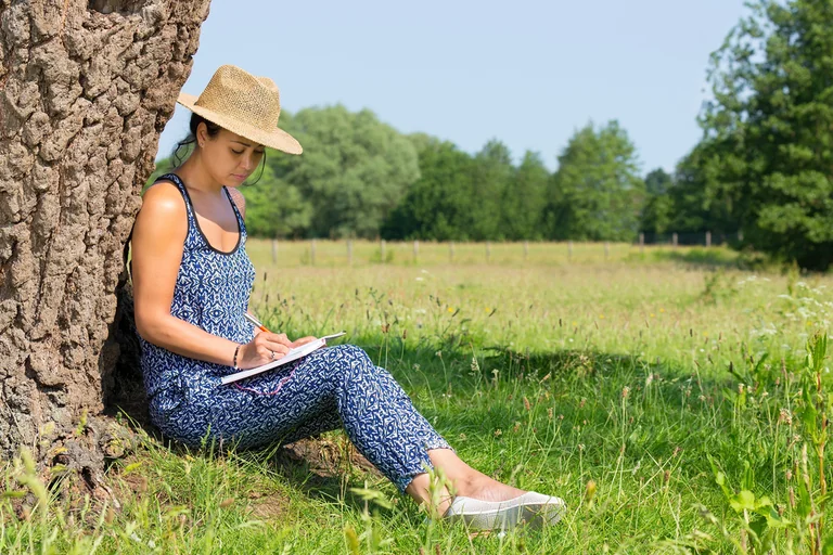 Frau mit Strohhut sitzt an einen Baum gelehnt auf einer Wiese und schreibt in ein Notizbuch
