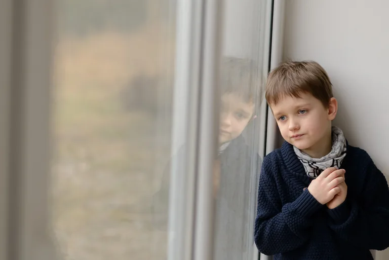 Nachdenklicher kleiner Junge in dunklem Pullover lehnt am Fenster und schaut hinaus