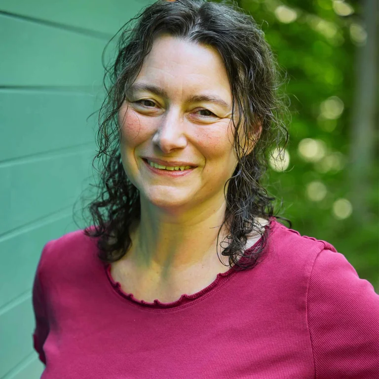 Sandra Quedenbaum, Coach für Hochsensible, lächelt freundlich in die Kamera, im Hintergrund eine grüne Holzwand und Bäume