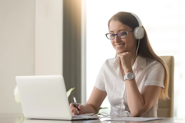 Frau mit Kopfhörer und Brille sitzt lächelnd am Laptop und macht sich Notizen