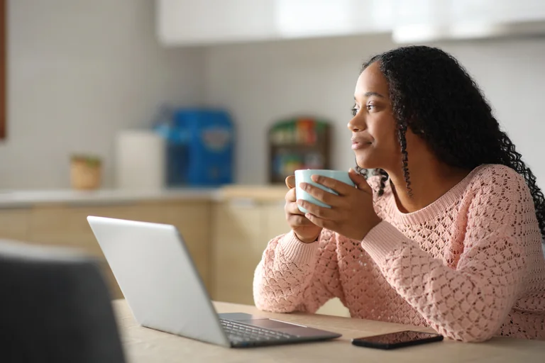 Zeit für Ruhe und Klarheit 🙂 Frau mit lockigem Haar hält eine hellblaue Tasse in beiden Händen und schaut nachdenklich aus dem Fenster, vor ihr ein Laptop