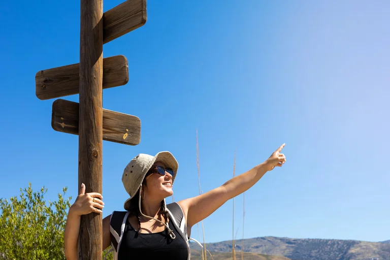 Wanderin mit Sonnenhut und Rucksack lehnt an einem hölzernen Wegweiser und zeigt mit ausgestrecktem Arm in die Ferne